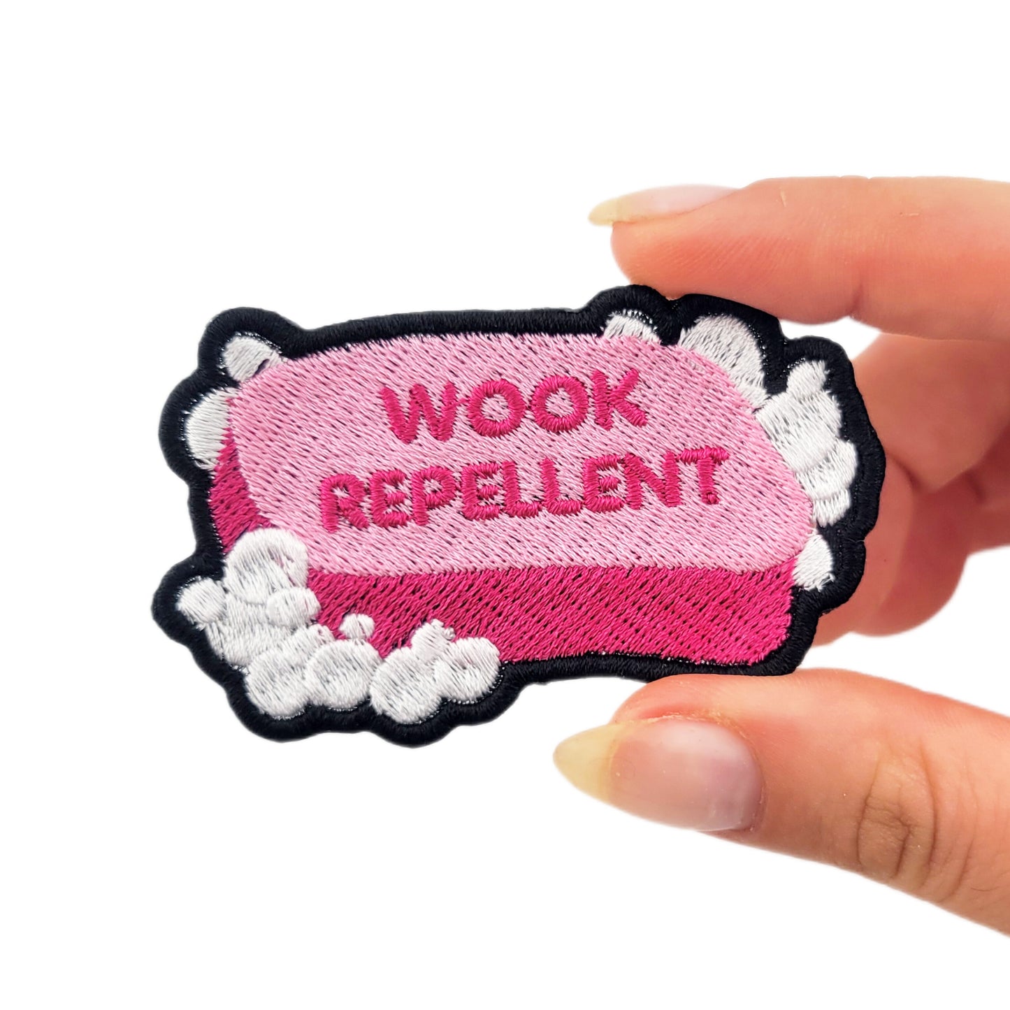 Patch with 'WOOK REPELLENT' text on a pink bar of soap, surrounded by white suds/bubbles. Held by a hand on a white background to demonstrate size.