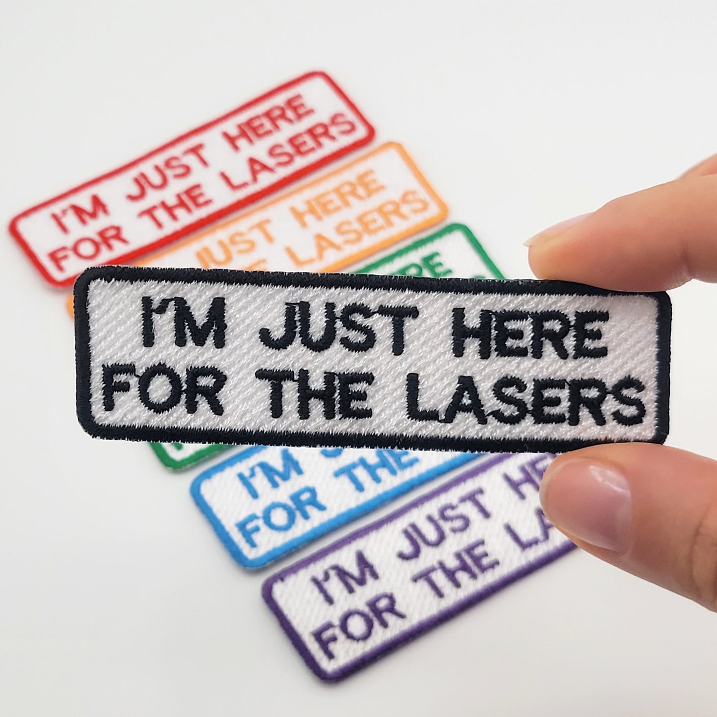 Embroidered patch with 'I'm just here for the lasers' text held by a hand, with other similar patches in the background.