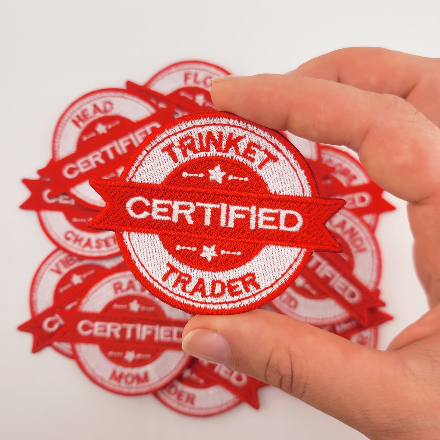 Red 'Certified Trinket Trader' patch held by a hand on a white background