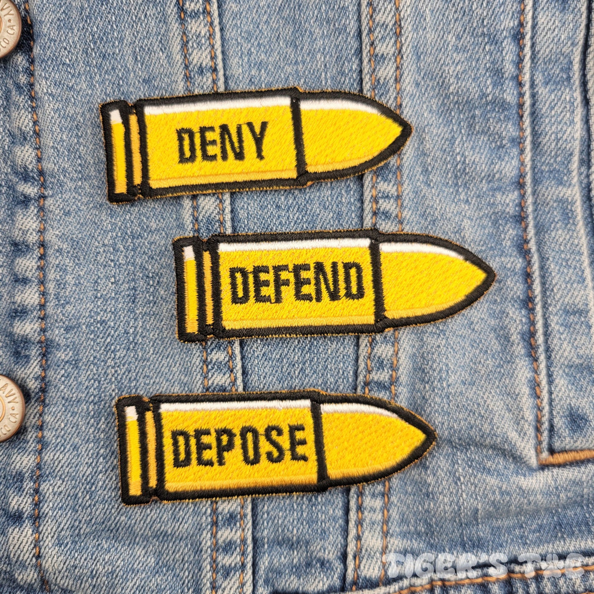 3 bullet-shaped yellow embroidered patches with the text "deny, defend, depose" written on each one, respectively. Displayed on a denim jacket background.