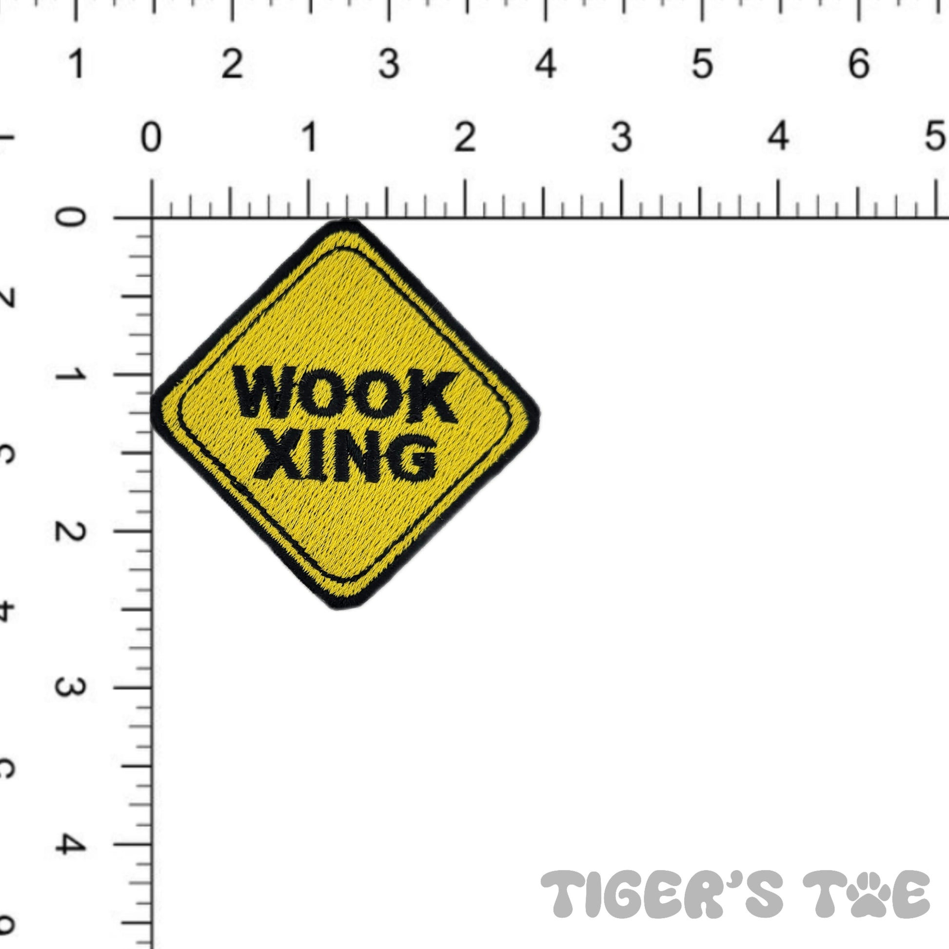 yellow embroidered patch in the shape of a crossing sign that reads 'wook xing' against a ruler for scale