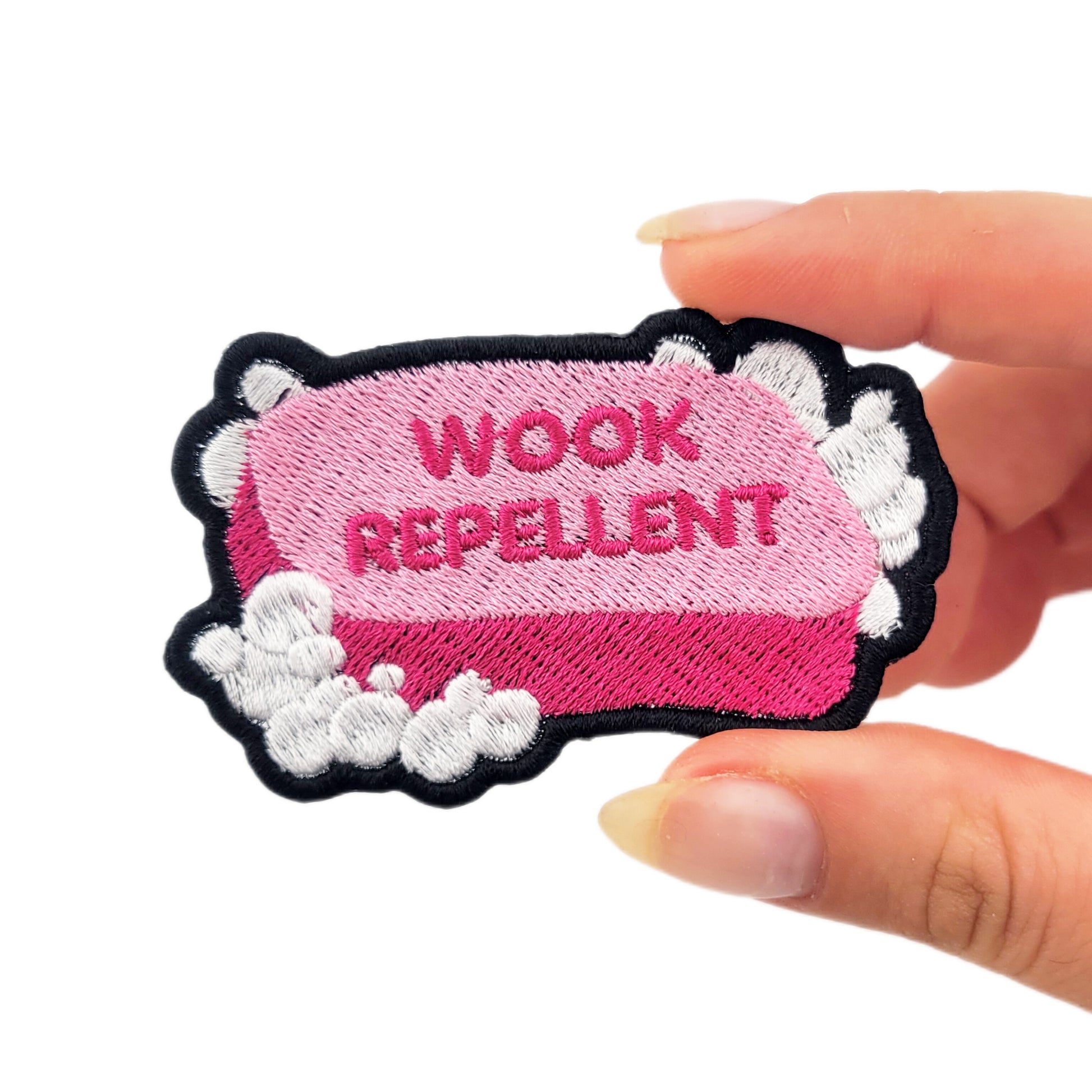 Patch with 'WOOK REPELLENT' text on a pink bar of soap, surrounded by white suds/bubbles. Held by a hand on a white background to demonstrate size.